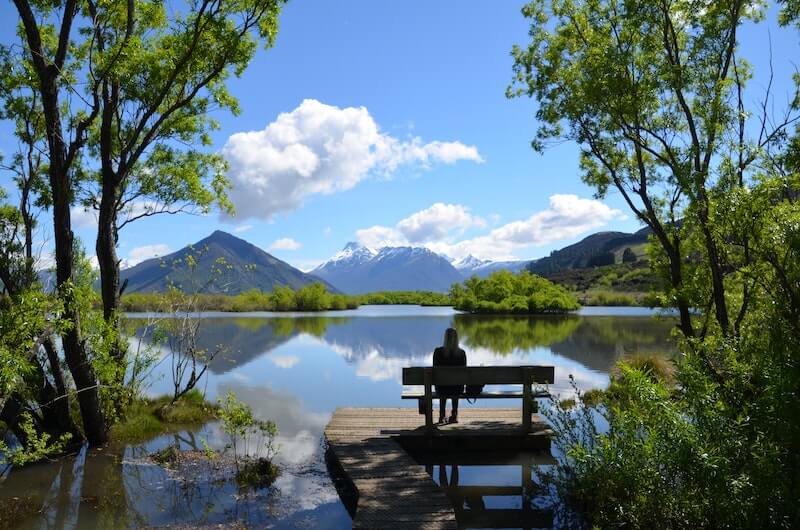 A woman sitting on a bench amongst trees at the end of a boardwalk leading to a pond with mountains in the background